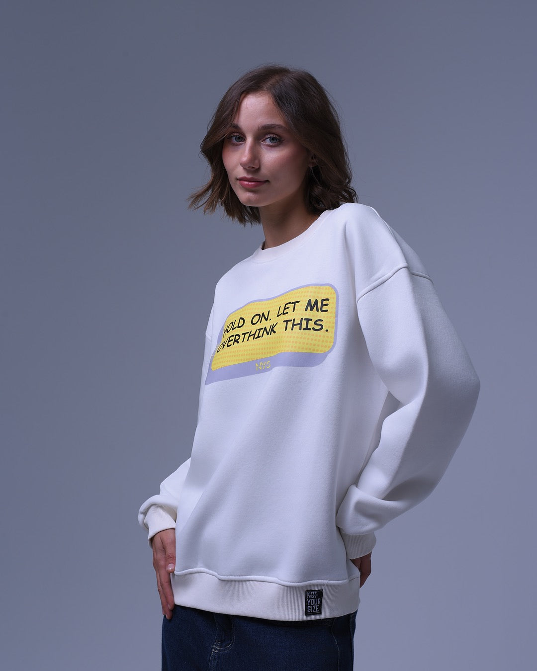 White oversized crewneck sweatshirt with yellow text graphic worn by woman posing against gray background